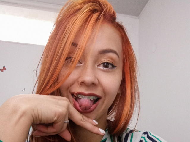 ElizaJones a les cheveux orange, elle tire sa langue avec son doigt, elle montre ses dents en métal et elle a du crayon noir pour les yeux. Elle sourit avec une expression mignonne et espiègle..