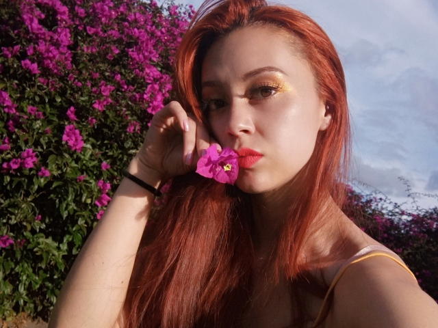 Eliza Jones, con cabello rojo, sombra de ojos dorada y una flor rosa en la boca, chupándola. Lleva una pulsera negra en la muñeca. La luz del sol ilumina su rostro..