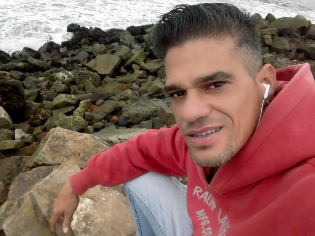 Supercockxx, con cabello corto y oscuro, una sudadera roja y auriculares blancos, sentado sobre rocas junto al agua, sonriendo ligeramente y mostrando un poco de sus dientes..