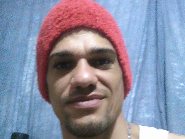 Supercockxx con un gorro rojo, sonriendo ligeramente, mostrando un rostro con barba incipiente, vistiendo una camiseta blanca, sobre un fondo de cortina azul..
