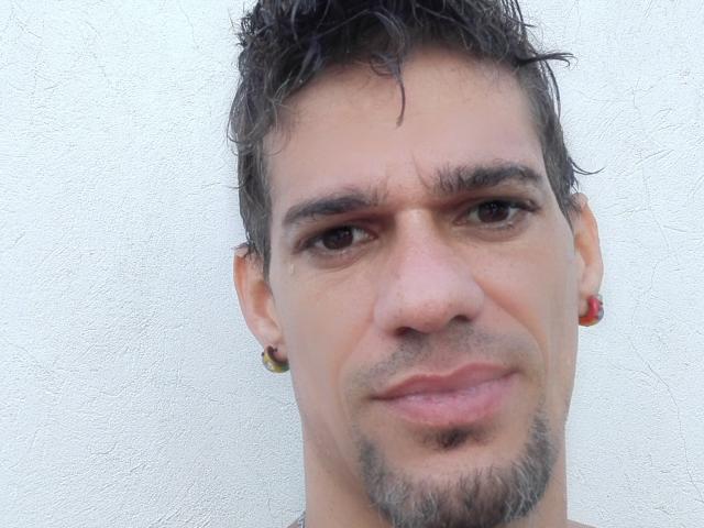 Supercockxx tiene el pelo corto, rizado y oscuro, pequeños pendientes rojos y una barba clara. Sus ojos marrones miran ligeramente hacia un lado y tiene una pequeña sonrisa discreta..