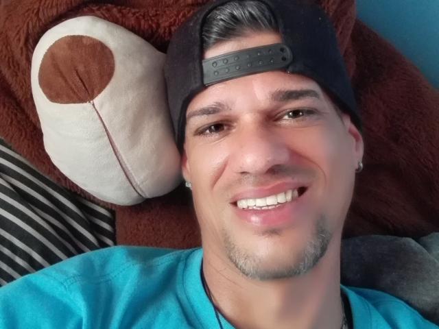 Supercockxx with a cute smile, wearing a black backward cap and a turquoise shirt, has a small patchy goatee and earrings, lying with a brown stuffed animal.