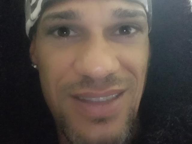 Ein Mann mit einer Mütze, braunen Augen, einer kleinen Nase, leichtem Bart und einem leichten Lächeln, bei dem seine Zähne sichtbar sind. Seine Haut ist hellbraun, und er hat einen kleinen Schnurrbart..