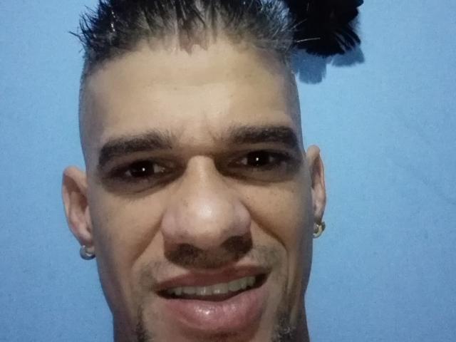 O Supercockxx tem um corte de cabelo curto e espetado com uma pena preta, um nariz pequeno e um sorriso discreto com os dentes à mostra. Ele está usando um pequeno brinco de argola na orelha esquerda..