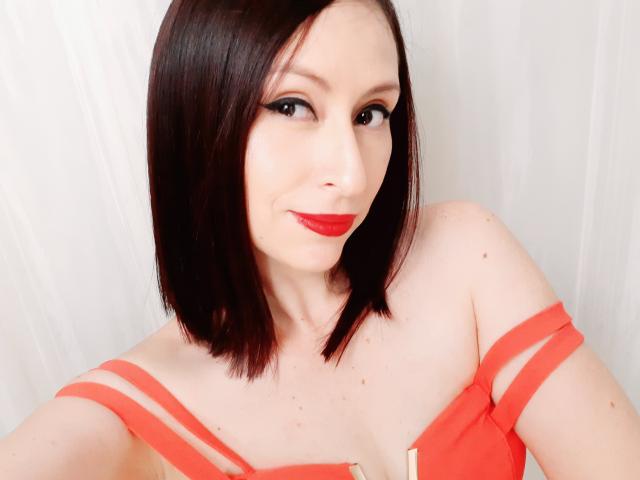 AliceRossi with shoulder-length black hair, red lipstick, and orange strap dress, showing her tits, smiling at the camera. Pale skin, dark eyeliner, white background.
