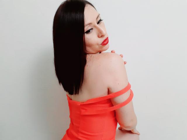 AliceRossi with shoulder-length black hair, red lipstick, and off-shoulder orange dress, looking over shoulder, showing her back and freckles, with hand near neck.