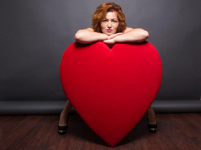 MelissaRilley est appuyée contre un énorme cœur rouge, les bras croisés, avec des cheveux roux bouclés, du rouge à lèvres rose et des talons hauts noirs, sur un plancher en bois..