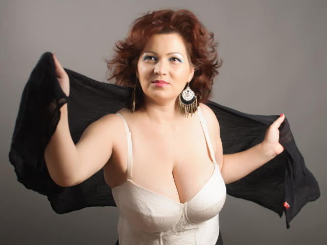 MelissaRilley met krullend rood haar, grote borsten in een witte top met kant, die een zwarte doek vasthoudt, met grote oorbellen, die iets opzij kijkt, in een studio..