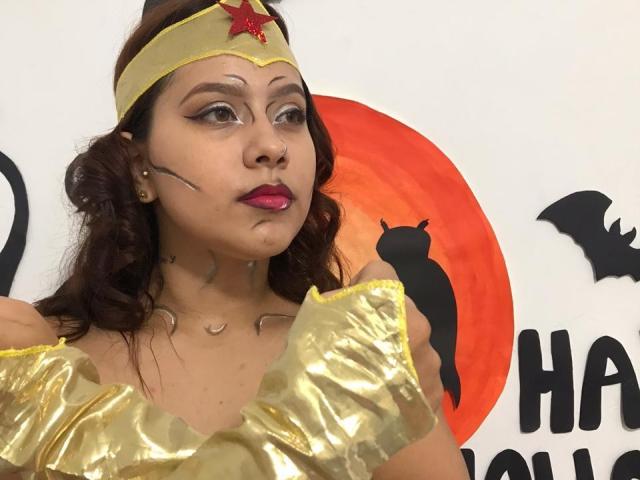 KorinYAmbar a les cheveux bruns et ondulés, porte un bandeau de super-héroïne jaune avec une étoile rouge, et une robe dorée brillante avec des manches froncées. Elle porte du maquillage sur son visage..