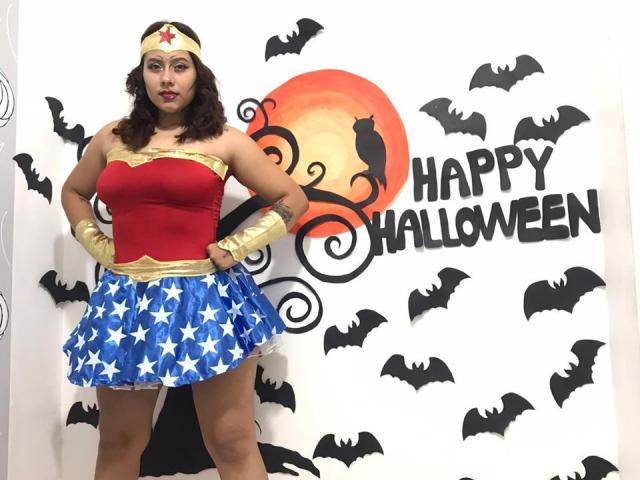 KorinYAmbar a les cheveux bruns bouclés, porte un haut rouge, une armure dorée, une jupe bleue en forme d'étoile et des bracelets dorés, et se tient là en cosplay de Wonder Woman, avec des silhouettes de chauves-souris et un coucher de soleil sur le mur..