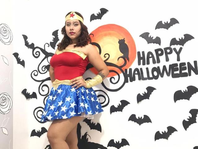 KorinYAmbar in a Wonder Woman costume with a red top, gold belt, blue skirt with stars, yellow gloves, and headband, standing with hands on hips, showing her ass.