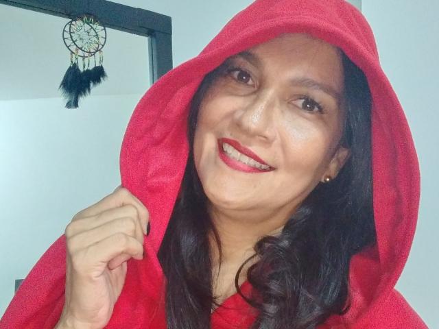 MatureJennyX with red hood, black hair, red lipstick, smiling, right fist clenched, wearing red hoodie, small earrings, dreamcatcher in mirror.