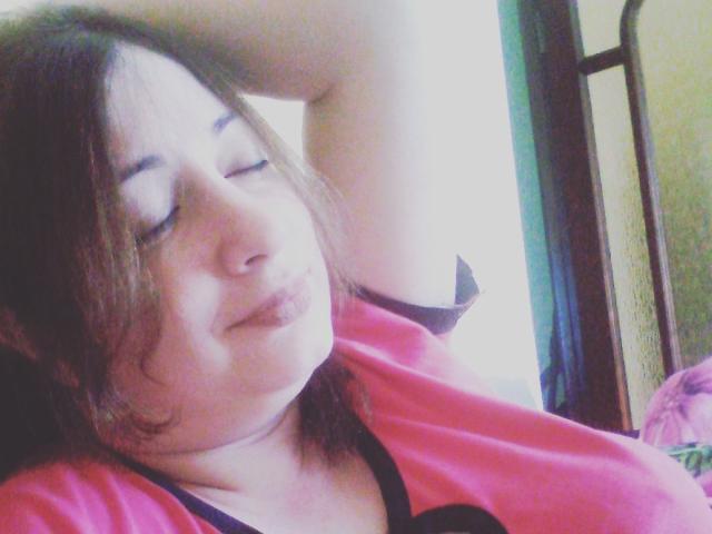 LediSusana, con labbra piene, capelli castani e una camicia rosa, occhi chiusi, braccio alzato, sorride leggermente, mostrando il sedere e i seni in primo piano..