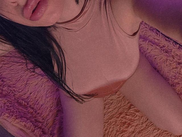 La canottiera rosa di MaxSandra, che mostra i suoi seni, con i capelli neri, le labbra rosa e la pelle illuminata di viola, è seduta su una soffice coperta rosa. Il suo inguine è leggermente visibile..