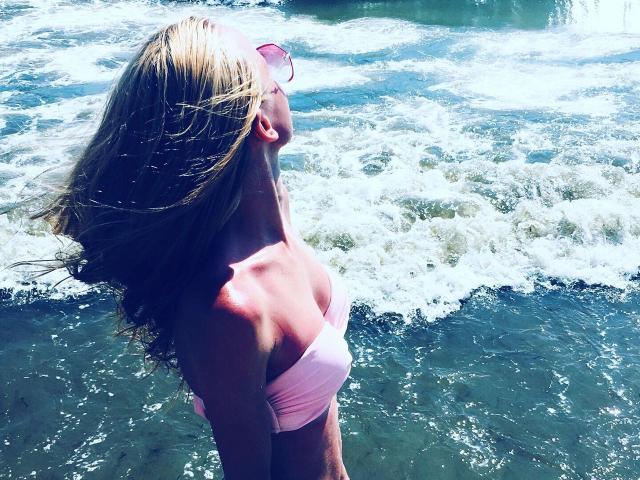 SusanSmite, con i capelli biondi, gli occhiali da sole rosa e un top rosa senza spalline, in piedi in acqua bassa, con la testa inclinata all'indietro, che guarda il cielo, con l'acqua che schizza intorno a lei..
