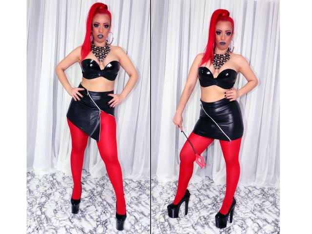 Diabelle, uma mulher com cabelo ruivo vibrante, usando um corpete preto sem alças, uma saia de couro preta, collant vermelhos, saltos altos pretos, segurando uma alça preta e usando um colar com espinhos..