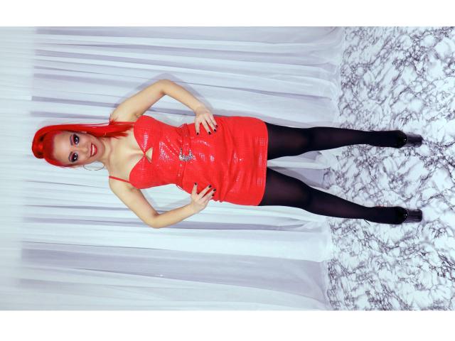 Diabelle, uma mulher com cabelo ruivo vibrante, vestindo um vestido de látex vermelho brilhante, collant preto e sapatos pretos, em pé com as mãos nos quadris, sorrindo para a câmera..