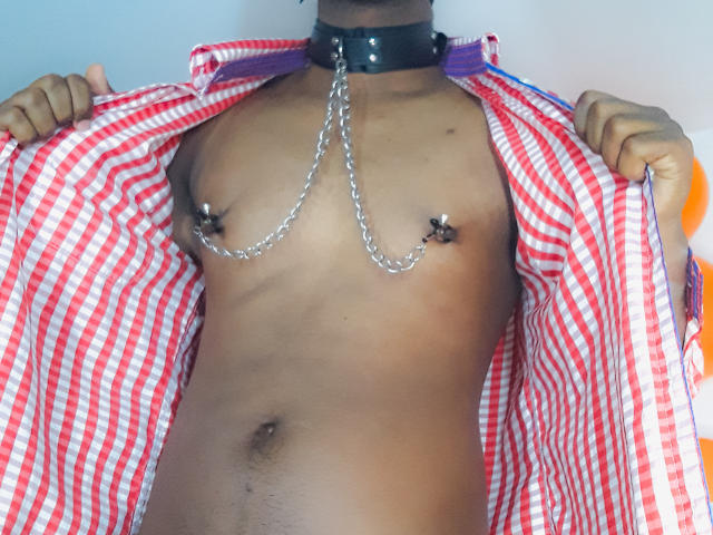MrJhon69 met donkere huid, met metalen ringen door zijn tepels, draagt een open rood-wit geruit shirt, een zwarte leren kraag, en geen broek, waarbij een plat, behaard borstgedeelte zichtbaar is..