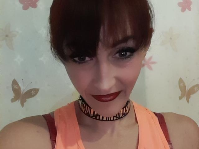 Xelenaxcbbs porte du rouge à lèvres, un maquillage des yeux foncé et un collier noir avec le mot "lucia" écrit en cursive. Elle porte un haut orange avec des bretelles rouges, et a les cheveux bruns..