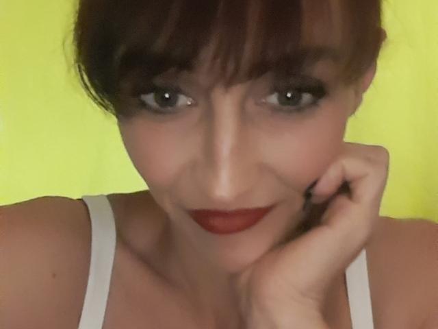 Xelenaxcbbs con rossetto rosso, trucco occhi scuro e canotta bianca, con il mento appoggiato sulla mano, sorride leggermente, capelli castani con frangia, sfondo giallo, occhi grigi, pelle chiara..