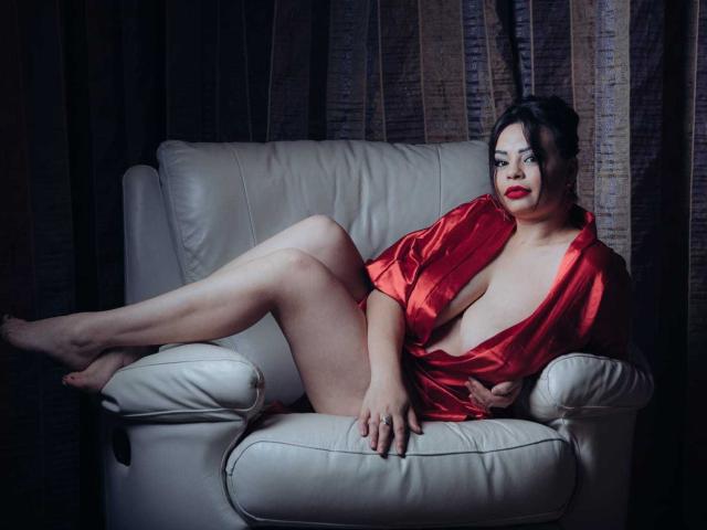 SexyHotSamira está recostada en una silla de cuero blanco, vistiendo una bata de satén rojo que revela sus grandes senos y su trasero desnudo. Tiene el pelo oscuro, lápiz labial rojo y una expresión segura..