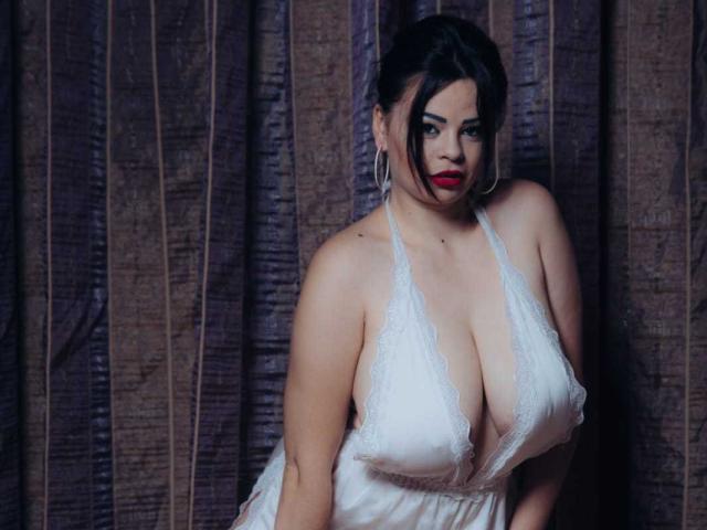 SexyHotSamira met grote borsten in een witte kanten halterjurk, rode lipstick, donker haar in een knot, met grote oorringen, staand voor een bruin gordijn..
