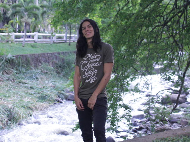 Antonio Santos, mit langem, schwarzem Haar, lächelnd, trägt ein grünes T-Shirt mit weißer Schrift, schwarze Hosen und steht neben einem plätschernden Bach, umgeben von Grün..