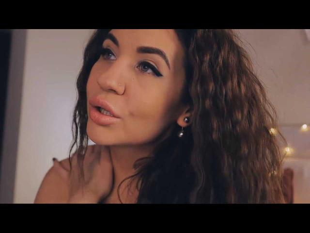 LolaSplash heeft krullend bruin haar, blauwe eyeliner en een neusring. Ze kijkt omhoog met lichtroze lippen, één hand in de buurt van haar nek. Haar borsten zijn niet zichtbaar..