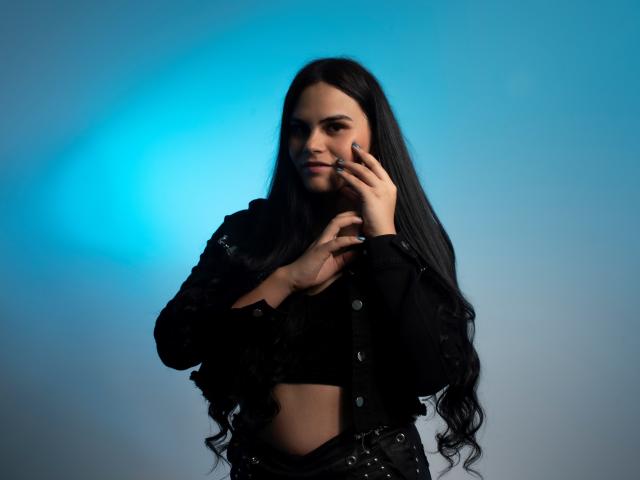 JassmineTS with long black hair, wearing a black crop top and jacket, touching her face with fingers, blue light background, serious expression, black nails, subtle makeup, slim figure.