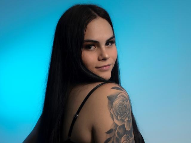 JassmineTS has long black hair, a rose tattoo on her right shoulder, and is looking over her shoulder with a slight smile, wearing a black bra. Blue background.
