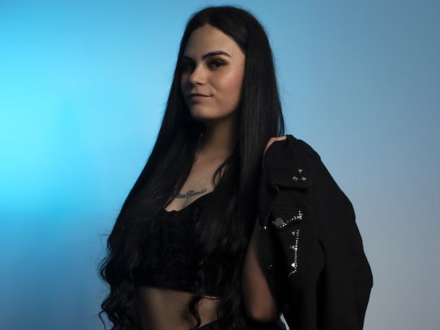 JassmineTS hat langes, schwarzes Haar, trägt ein schwarzes Crop-Top und eine schwarze Jacke mit silbernen Nieten und zeigt ein Tattoo auf ihrer Brust. Der Hintergrund ist blau, und sie blickt leicht nach links..