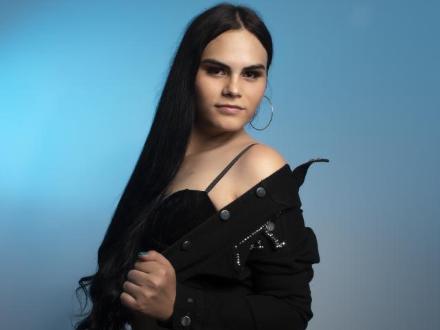 JassmineTS has long black hair, a confident look, and is wearing a black jacket with buttons, showing black straps, and large hoop earrings, against a blue background.