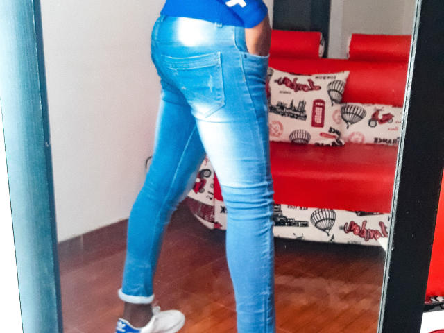 Le jean bleu moulant de MrJhon69 révèle un postérieur brillant et des jambes fines, avec des baskets blanches, dans un reflet miroir. Un canapé rouge avec un coussin à motifs est visible en arrière-plan..