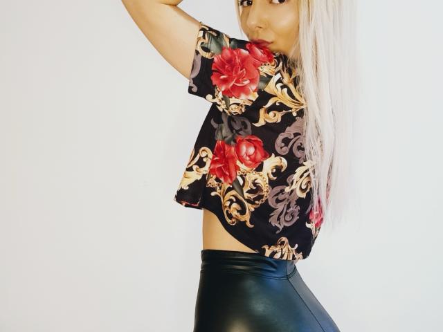 JuliCandy, met lang blond haar, draagt een zwarte crop top met bloemenprint en zwarte leren broek, waarbij haar buik zichtbaar is en ze haar arm omhoog heeft, terwijl ze over haar schouder kijkt..
