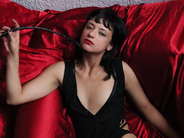 Marlaa, con cabello negro, lápiz labial rojo y un vestido negro elegante, sosteniendo un látigo de cuero negro sobre almohadones de satén rojo brillante, mirando seductoramente a la cámara..
