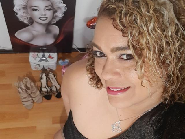 SolmaryHot with curly blonde hair, red lipstick, black lace top, and butterfly necklace, smiling at camera, standing next to Marilyn Monroe poster, and various high heels on wooden floor.