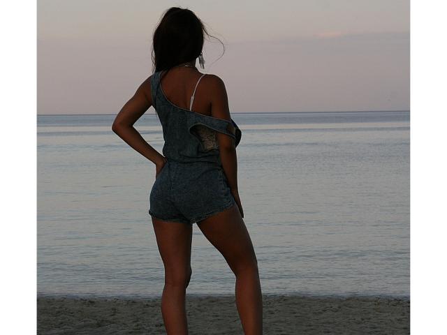 Amelia steht am Strand, dem Ozean zugewandt, und trägt ein lockeres, graues Oberteil, das von einer Schulter fällt und einen weißen BH-Träger freilegt. Sie hat die Hände in die Hüften gestemmt und zeigt ihr Gesäß..