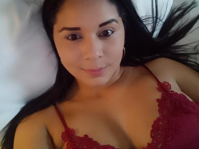 EvaMonn deitada em uma cama, vestindo um sutiã de renda vermelho, mostrando seus seios, com cabelos longos e pretos, olhos castanhos e um leve sorriso. Iluminação suave..