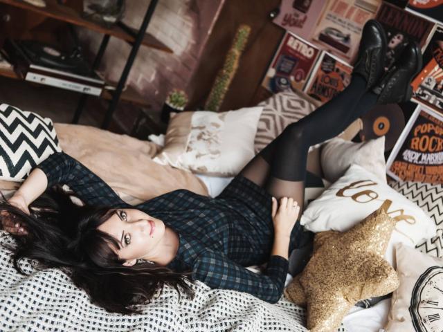 MissGulia is lying on a bed with her legs up, wearing a dark plaid dress, black stockings, and black boots, her tits slightly visible, dark brown hair, and a smile.
