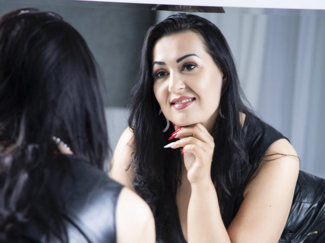 Linda, les cheveux noirs, souriant dans un miroir, portant un haut noir sans manches, se tenant le menton, avec des ongles rouges, ayant l'air mignonne, dans une pièce lumineuse..