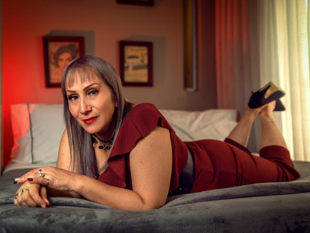 DianeOasis, sdraiata su un letto, indossa un vestito rosso, una collana nera e dei tacchi neri, con i glutei in evidenza, guarda la fotocamera, sorride e mostra il seno..