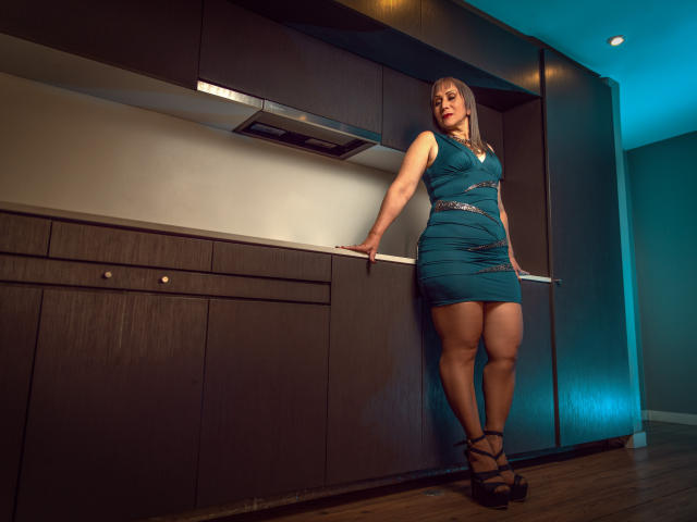 DianeOasis stands in a kitchen, leaning on dark brown cabinets. She wears a teal dress, black high heels, and has a curvy, thick-figured body with big ass and full tits.