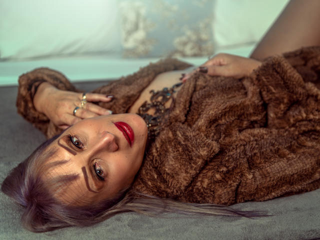 DianeOasis with purple hair, red lipstick, and brown fur coat, lying on grey fabric, hand on chest, black necklace, looking at camera, confident, sexy.
