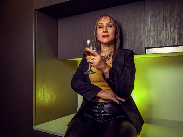 DianeOasis with brown hair, red lipstick, black pinstripe blazer, yellow top, black leather pants, holding glass of red wine, sitting, green-lit background, confident, sexy, mature.