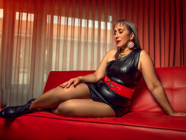 DianeOasis, mit ihrem Hintern auf einer roten Ledercouch sitzend, trägt ein schwarzes Latexdress, einen roten Gürtel, schwarze Stiefel und pinke Ohrringe, und wirkt dabei sexy und selbstbewusst..