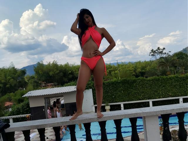 LindaAfrodita in un bikini rosso, in piedi su un balcone con una mano tra i capelli, che mostra il suo sedere tonico e i suoi piccoli seni, con una piscina e vegetazione sullo sfondo..