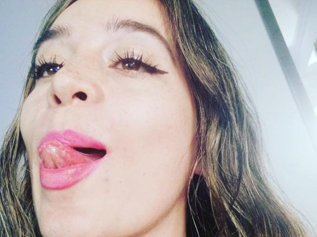Het gezicht van Valentina Marina, met roze lippenstift, haar tong uitgestoken, lange wimpers en golvend bruin haar. Haar uitdrukking is speels en ze heeft een lichte glimlach. Haar borsten zijn niet zichtbaar..
