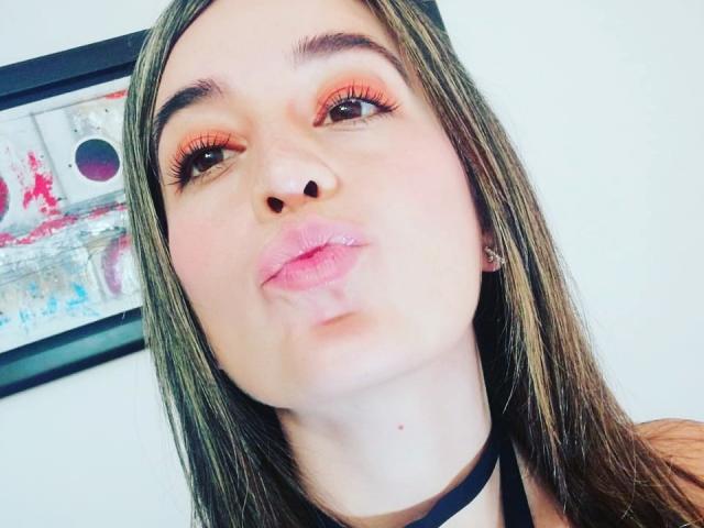 Valentina Marina, con labbra rosa carnose, occhi marroni e ombretto arancione, fa una smorfia come per dare un bacio. Ha lunghi capelli castani, una collana nera e un nasino carino..