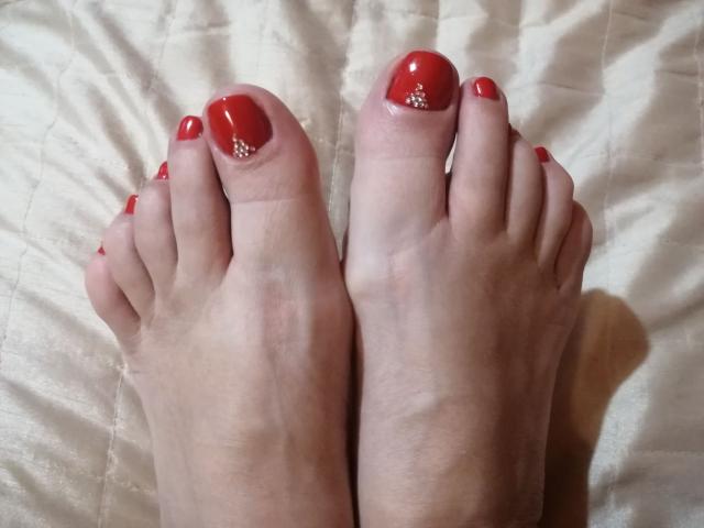 Los pies de LadyEvelynn, con esmalte de uñas rojo brillante, pequeños puntos brillantes en los dedos grandes, y piel pálida, colocados uno al lado del otro sobre una colcha blanca con textura..