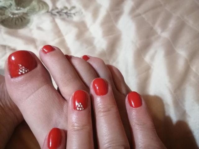 De rode nagels van LadyEvelynn, met glanzende nagellak en kleine witte glitters op de ringvinger en middelvinger, handen rustend tegen elkaar op een crème-kleurige stof..